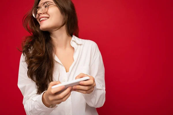 흰색 셔츠와 광학 안경을 착용하고 빨간 배경 위에 고립 된 예쁘게 웃고 있는 젊은 브라 무네 여성의 사진, 손에 핸드폰을 들고 온라인 게임을 하고 있는 모습 — 스톡 사진