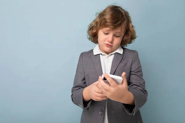 Foto von hübschen kleinen männlichen Kind mit lockigen Haaren trägt grauen Anzug halten und mit Handy isoliert über blauem Hintergrund Blick auf Smartphone kommunizieren vie sms — Stockfoto
