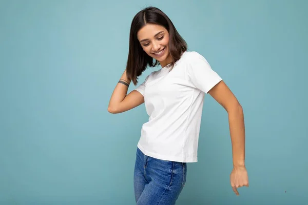 肯定的な笑顔の喜び若い美しいブルネットの女性で誠実な感情を身に着けていますカジュアルホワイトTシャツのためにモックアップのために隔離された青の背景上に空のスペースとダンス — ストック写真