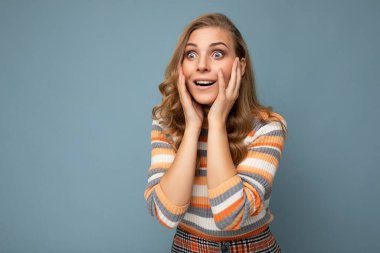 Genç, güzel, sarışın, kıvırcık bir kadının fotoğrafı. Samimi duyguları olan, sıradan çizgili kazak giyen. Mavi arka planda izole edilmiş, boş alan olan.