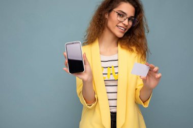 Sarı ceketli ve optik gözlüklü güzel bir kadının portresi arka plandaki duvarda izole edilmiş ve boş ekran ve kredi kartıyla kameraya bakan telefonu gösteriyor.