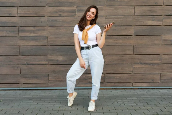 Full length body size photo of pretty smiling female person wearing casual clothes standing in the street holding and using mobile phone looking at camera — Stock Photo, Image