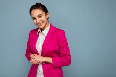 Çekici, güzel, pozitif gülümseyen esmer kadının portresi samimi duyguları olan, günlük beyaz gömlek giyen ve mavi arka planda izole edilmiş, modaya uygun pembe ceket giyen.