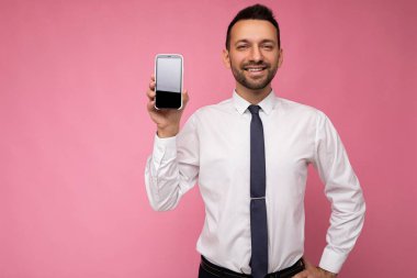 Yakışıklı, yakışıklı, yakışıklı, şık kıyafetler giyen, renkli arka plan duvarında tek başına duran, akıllı telefon tutan ve boş ekran ekranlı telefonu gösteren yakışıklı bir adam.