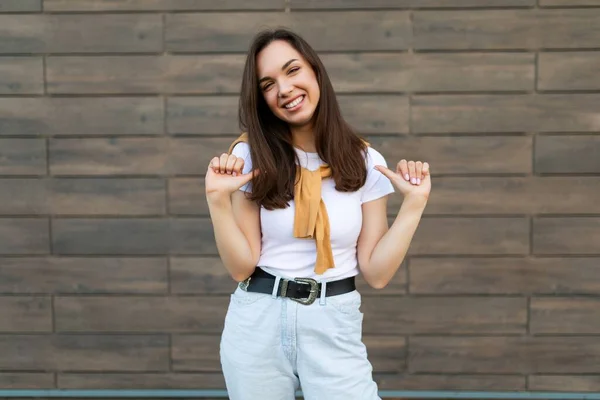 Portrait of young beautiful positive happy brunette woman standing against brown wall in the street and wearing stylish outfit — Stock Photo, Image