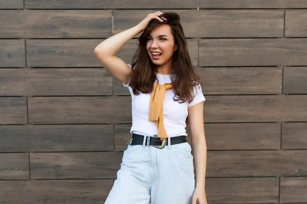 Porträtt av framgångsrika leende glad glad ung brunett kvinna bär casual vit t-shirt och jeans med gul tröja balanserar nära brun vägg på gatan och har kul — Stockfoto