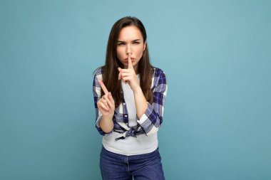 Samimi duyguları olan, mavi arka planda izole ve sessiz bir jest yapan, modaya uygun bir çek tişörtü giyen genç, sinirli, güzel bir esmer kadın. Gizli kavram tut