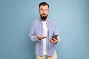 Yakışıklı, esmer, sakalsız, şık beyaz tişörtlü ve mavi gömlekli bir adam. Mavi arka planda izole edilmiş. Elinde boş bir alan var.