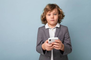 Kıvırcık saçlı, takım elbiseli, mavi arka planda izole edilmiş bir telefonu olan yakışıklı çocuk kameraya bakıyor.