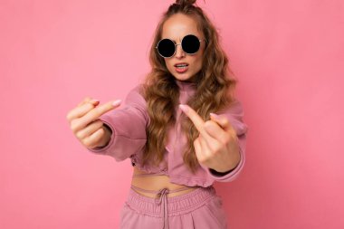 Kendine güveni tam, güzel, sarışın, samimi duyguları olan şık, pembe spor takım elbiseli ve pembe arka planda izole edilmiş gözlüklü orta parmaklı bir kadın.