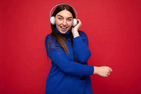Attractive positive young brunette woman wearing blue sweater isolated over red background wall wearing white bluetooth earphones listening to cool music and having fun looking at camera