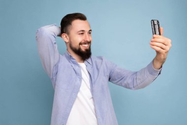 Yakışıklı, esmer, sakallı, sıradan kıyafetler giyen, mavi arka plan duvarında izole bir şekilde duran akıllı telefon taşıyan, cep telefonu ekran ekranına bakarak selfie çeken bir adam.