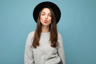 Genç, güzel esmer kadın. Gri kazaklı ve şık siyah şapkalı modaya uygun bir bayan. Pozitif kadın yüz samimi duygularını gösterir. Mavi arkaplanda izole edilmiş, boş alan veren komik bir model