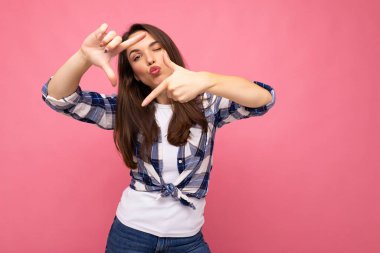 Genç, pozitif, hoş, gülümseyen, samimi duyguları olan, hippi tişörtü giyen, ellerle çerçeve yapan, hayali kamerayla fotoğraf çeken ve pembe üzerine izole edilmiş güzel esmer kadın.