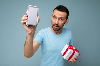 Yakışıklı, esmer, sakallı, mavi arka plan duvarında sakallı, mavi tişörtlü, elinde kırmızı kurdeleli, cep telefonlu beyaz bir hediye kutusu olan genç bir adam.