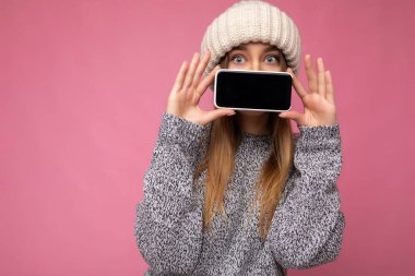 Kapalı çekimde, pembe arka planda izole edilmiş gri süveter ve bej şapka giyen güzel sarışın bir kadının el ele tutuşmuş ve cep telefonunu boş bir şekilde gösteren bir fotoğraf.