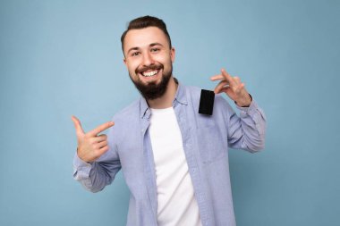 Güzel görünüşlü, sırıtan esmer, sakallı, şık mavi tişörtlü ve beyaz tişörtlü genç bir adam. Mavi arka plan duvarında izole edilmiş, elinde kredi kartıyla kameraya bakıyor ve parmağını işaret ediyor.