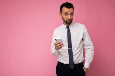 Düşünceli yakışıklı genç adam arka plan duvarından izole edilmiş günlük giysiler giyiyor ve cep telefonu yazıları yazıyor kameraya bakıyor ve düşünüyor.