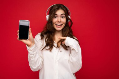 Çekici, pozitif, kıvırcık saçlı, beyaz gömlek giyen, kırmızı arka plan duvarında izole edilmiş, cep telefonlu, beyaz kulaklık takan, internette gezen,