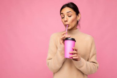Charming young happy brunette woman wearing stylish clothes isolated over colourful background wall holding paper cup for mockup drinking milkshake enjoying with close eyes