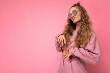 Attractive funny amusing young blonde woman wearing everyday stylish clothes and modern sunglasses isolated on colorful background wall looking to the side