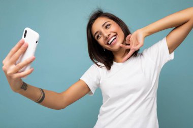 Çekici, genç, gülümseyen, mutlu bir kadın cep telefonu tutuyor ve kullanıyor. Selfie çekiyor. Duvar arkasından izole edilmiş şık kıyafetler giyiyor.