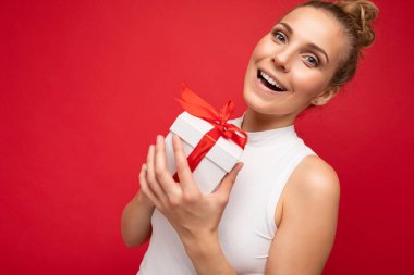Hoş, güzel, pozitif gülümseyen, şaşırmış, kırmızı arka plan duvarında izole edilmiş, beyaz bluzlu, elinde hediye kutusu olan ve kameraya bakan genç, sarışın kadın.