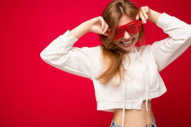 Renkli arka planda izole edilmiş, beyaz kapüşonlu ve şık güneş gözlüklü, güzel, mutlu, genç, sarışın bir kadın.