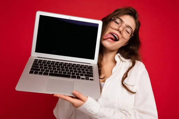 Crazy mad beautiful brunette curly young woman holding computer laptop wearing glasses white shirt showing tongue looking to the side isolated over red wall background