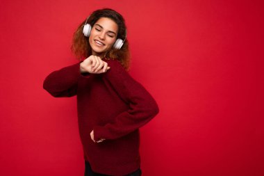 Çekici, pozitif, kıvırcık saçlı, koyu kırmızı süveter giyen, kırmızı arka plan duvarında izole edilmiş, beyaz bluetooth kulaklık takan, havalı müzik dinleyen, eğlenen ve hareket eden bir kadın.