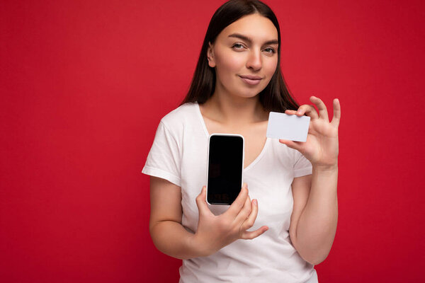 Фото красивой юной брюнетки в белой футболке на красном фоне с кредиткой и мобильным телефоном с пустым дисплеем, смотрящим в камеру