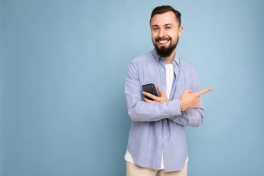 Gülümseyen, genç, esmer, sakalsız, şık beyaz tişörtlü ve mavi gömlekli bir adam. Mavi arka planda, elinde boş bir alanda kameraya bakıyor.