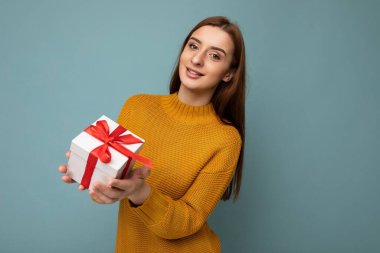Güzel, mutlu, genç esmer kadın renkli arka plan duvarında izole edilmiş şık günlük kıyafetler giymiş elinde hediye kutusuyla kameraya bakıyor.