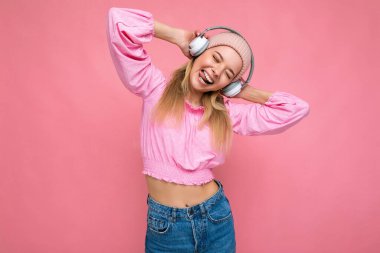 Çekici, pozitif gülümseyen, pembe bluz giyen, pembe şapkalı, arka plan duvarına izole edilmiş, beyaz kablosuz kulaklık takan, müzik dinleyen genç, sarışın bir kadının fotoğrafı.