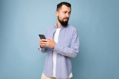 Düşünceli yakışıklı genç adam arka plan duvarından izole edilmiş günlük giysiler giyiyor ve cep telefonu yazıları kullanıyor.