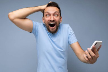 Aşırı duygusal, yakışıklı, sakallı, esmer adam günlük mavi tişört giyiyor. Mavi arka plan üzerinde izole edilmiş. İnternette cep telefonu iletişimini kullanıyor ve kameraya bakıyor.