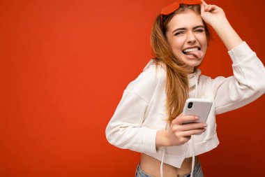 Güzel, duygusal, genç, sarışın bir kadın şık beyaz kapüşonlu ve komik renkli gözlükler takıyor. Renkli arka planda izole bir şekilde duruyor ve cep telefonuyla internette iletişim kuruyor.