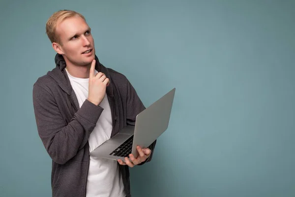 Side profile photo shot of handsome thoughtful blonde man holding computer laptop posing finger near chin wearing white t-shirt and grey sweater looking to the side isolated over blue background