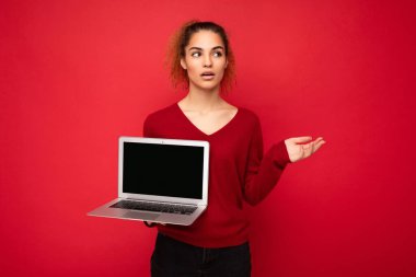 Güzel, kıvırcık, esmer, kırmızı kazaklı bir kadın kırmızı duvar arkasında izole bir şekilde duruyor. Elinde bilgisayarında boş fotokopi alanı olan bir bilgisayar var. Yan tarafı göstererek yan tarafa bakıyor.