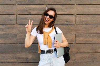 Güzel, gülümseyen, genç bir esmer kadın elinde bilgisayar dizüstü bilgisayar ve güneş gözlüğü ile sokakta açık mavi kot pantolon ve beyaz tişörtle barış jesti gösteriyor.