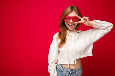 Güzel, pozitif sarışın bir kadının günlük kıyafetler giydiği ve renkli arka plan duvarından izole edilmiş gözlüklü bir şekilde kameraya bakarken ve barış jesti ve dil gösterirken çekilmiş bir fotoğrafı.