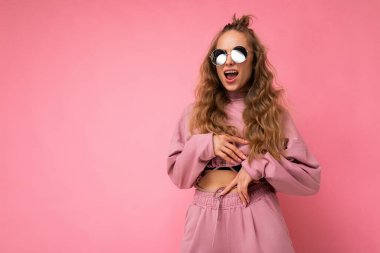 Güzel, mutlu, esmer, kıvırcık saçlı bir kadının pembe arka plan duvarından izole edilmiş, günlük pembe spor kıyafetleri ve şık güneş gözlükleriyle kameraya bakıp eğlendiği bir fotoğraf.