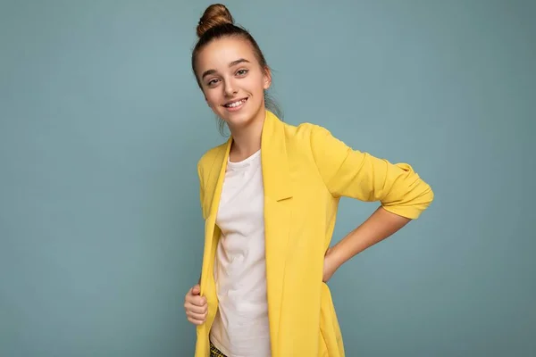 Foto de encantadora positiva feliz sonriente morenita pequeña adolescente con chaqueta amarilla de moda y camiseta blanca de pie aislado sobre la pared de fondo azul mirando a la cámara — Foto de Stock