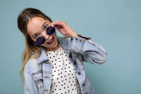 Fotografía de una joven rubia alegre bastante positiva con chaqueta de jean azul y gafas de sol elegantes aisladas sobre una pared de fondo azul mirando a la cámara — Foto de Stock