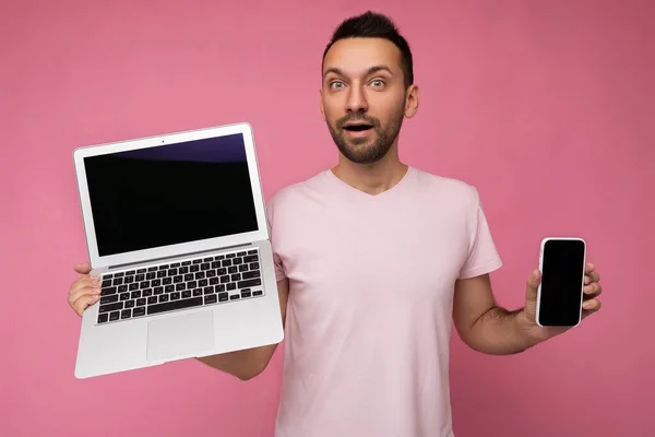 英俊而惊奇的男人拿着手提电脑和手机，穿着孤立的粉色背景T恤，看着相机 — 图库照片