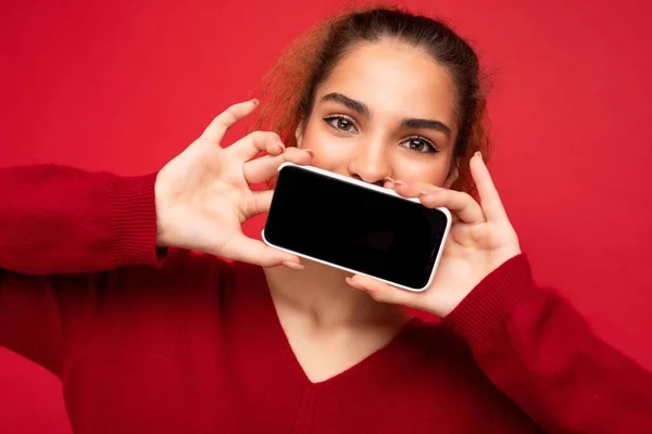 빨간 스웨터를 입고 빨간 배경에 고립된 젊은 여성 이 스마트폰을 들고 있는 모습 과 카메라를 볼 수있는 사본 공간 이 있는 휴대 전화 화면을 보여 주는 사진. — 스톡 사진