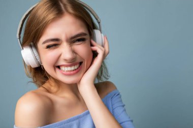 Çekici, mutlu, gülümseyen sarışın bir kadının portresi mavi arka planda izole edilmiş, beyaz kablosuz kulaklık takıyor, havalı müzik dinliyor ve eğleniyor.