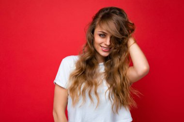 Beyaz tişört giyen güzel sarışın kadın kameraya bakıyor. Pozitif kadın yüz samimi duygularını gösterir. Boş alanı olan kırmızı duvarda izole edilmiş komik bir model