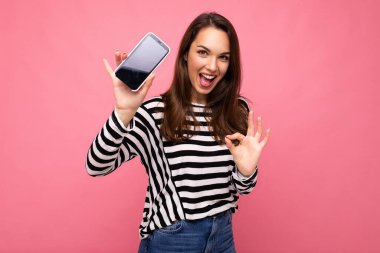 Güzel, mutlu, çizgili süveter giyen genç bir kadın. Arka planda izole edilmiş. Fotokopi alanı, cep telefonu ekranını gösteren kameraya iyi bir jest gösteriyor.