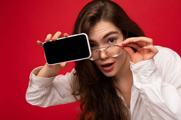 흰색 셔츠와 광학 안경을 착용 한 아름다운 젊은 갈색 여성 이 손에 붉은 색 배경에 고립되어 카메라를 보기 위해 빈 화면을 가진 휴대 전화를 보여 주고 있다. — 스톡 사진